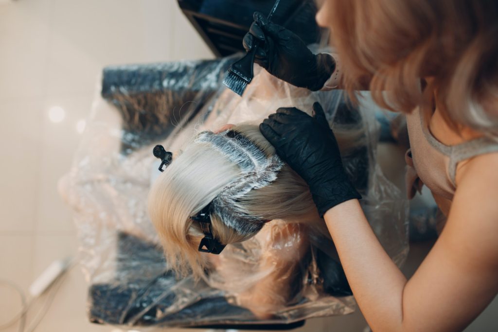 Blonde haired woman in a salon getting dark roots bleached by a stylist with black gloves