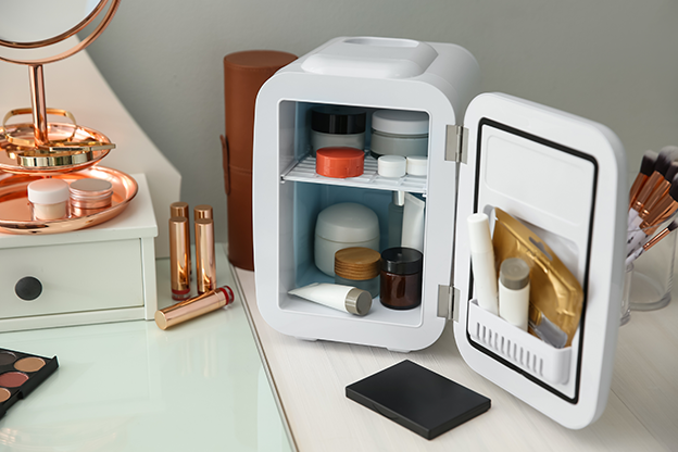 Mini skincare fridge with organized makeup and beauty products, placed on a vanity table alongside a makeup organizer, mirror, and cosmetics.