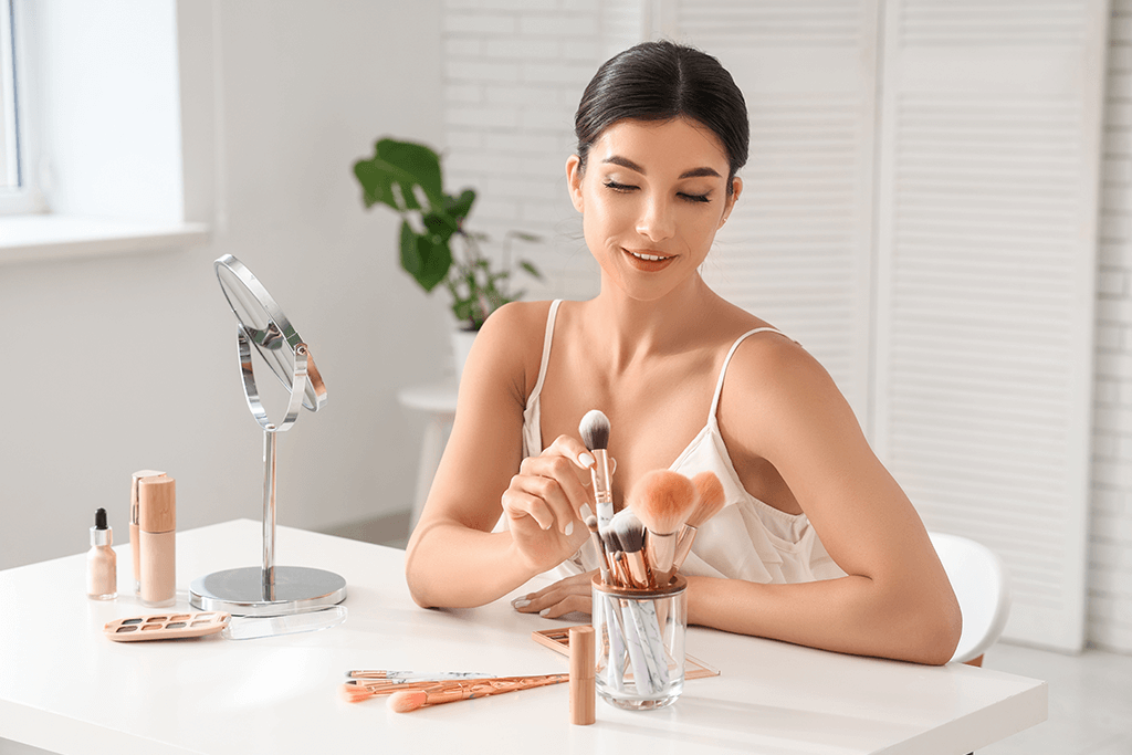 Smiling woman sitting at a white desk with a makeup organizer, brushes, mirror, and cosmetics, applying makeup in a bright and minimalist setting.