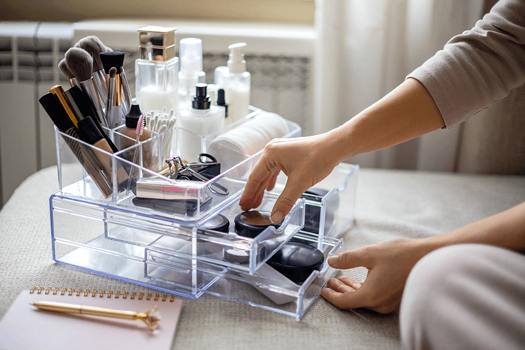 Clear acrylic makeup beauty organizer holding brushes, cosmetics, and skincare products, with a hand opening one of the drawers on a well-lit table.