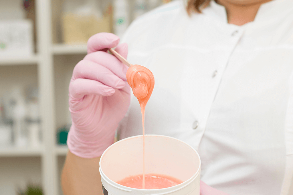 A beautician wearing pink gloves holding a spatula with warm wax, showcasing the preparation process for a professional waxing treatment.