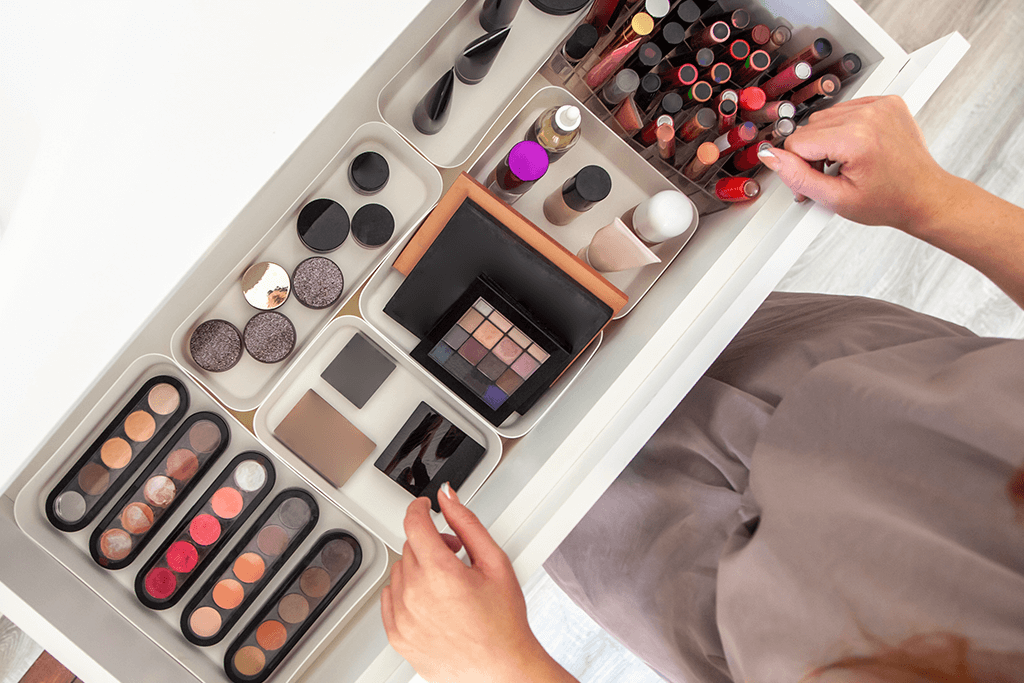 Clear acrylic makeup organizer holding brushes, cosmetics, and skincare products, with a hand opening one of the drawers on a well-lit table.