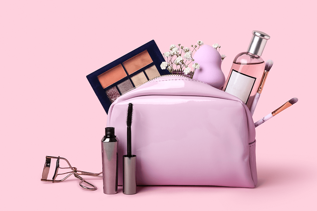 The image features a pink makeup bag with various beauty products and accessories spilling out. Items visible include an eyeshadow palette, mascara, eyelash curler, a perfume bottle, a beauty blender sponge, and several makeup brushes, all arranged on a soft pink background. The combination of cosmetics, tools, and flowers suggests a fresh, stylish beauty setup.